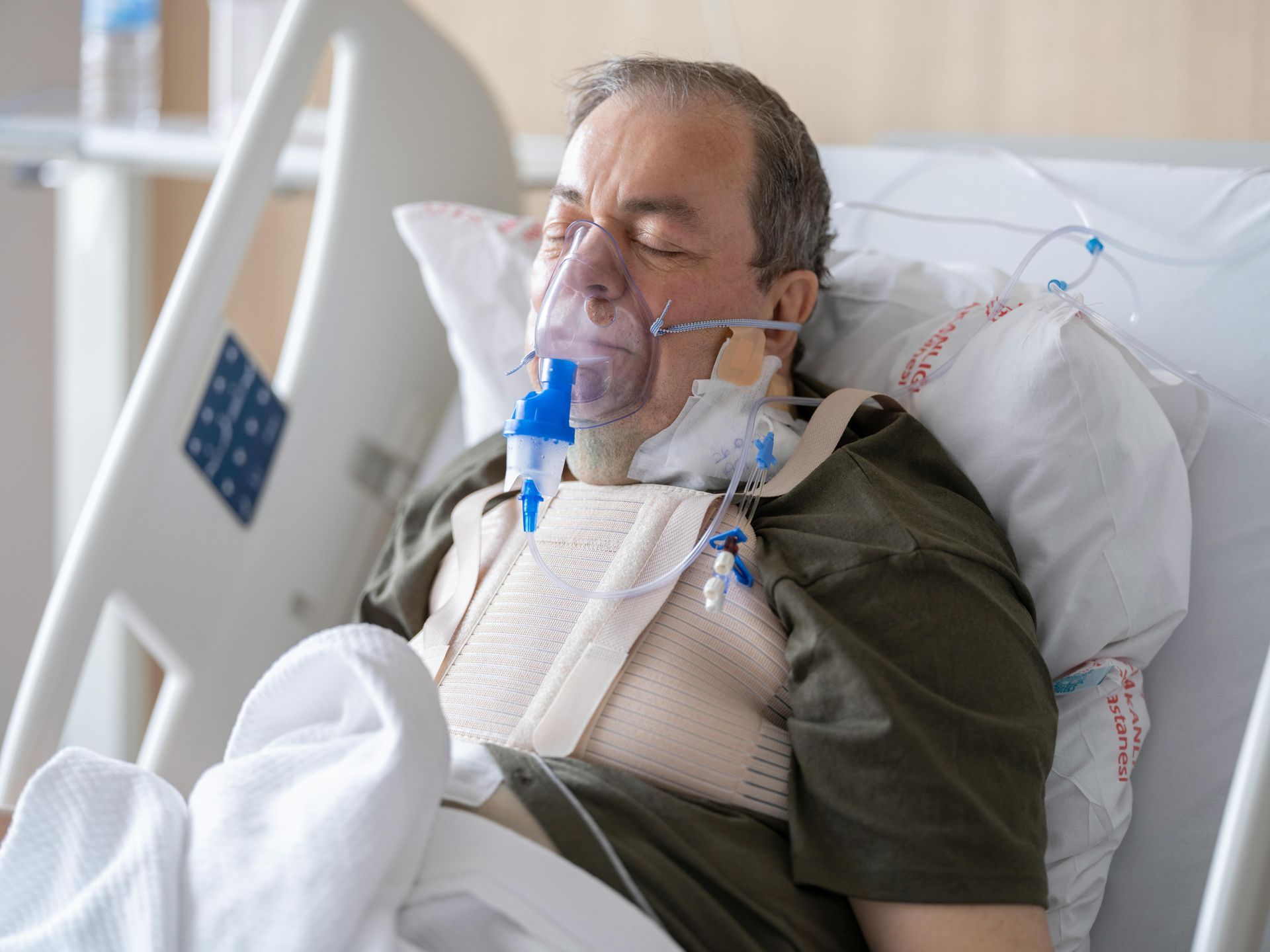 Man in hospital bed with oxygen mask and chest support.