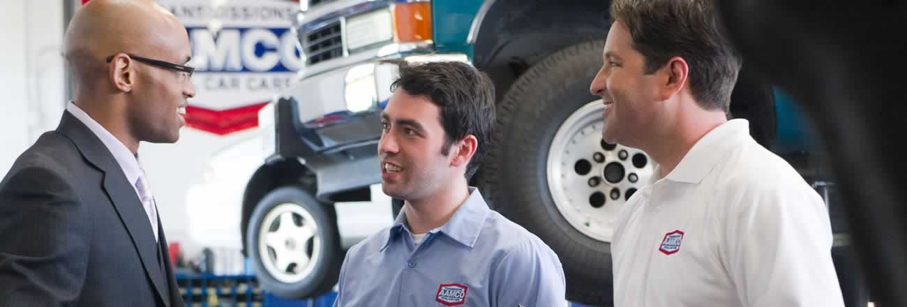 2 AAMCO techs with customer in front of vehicle on lift