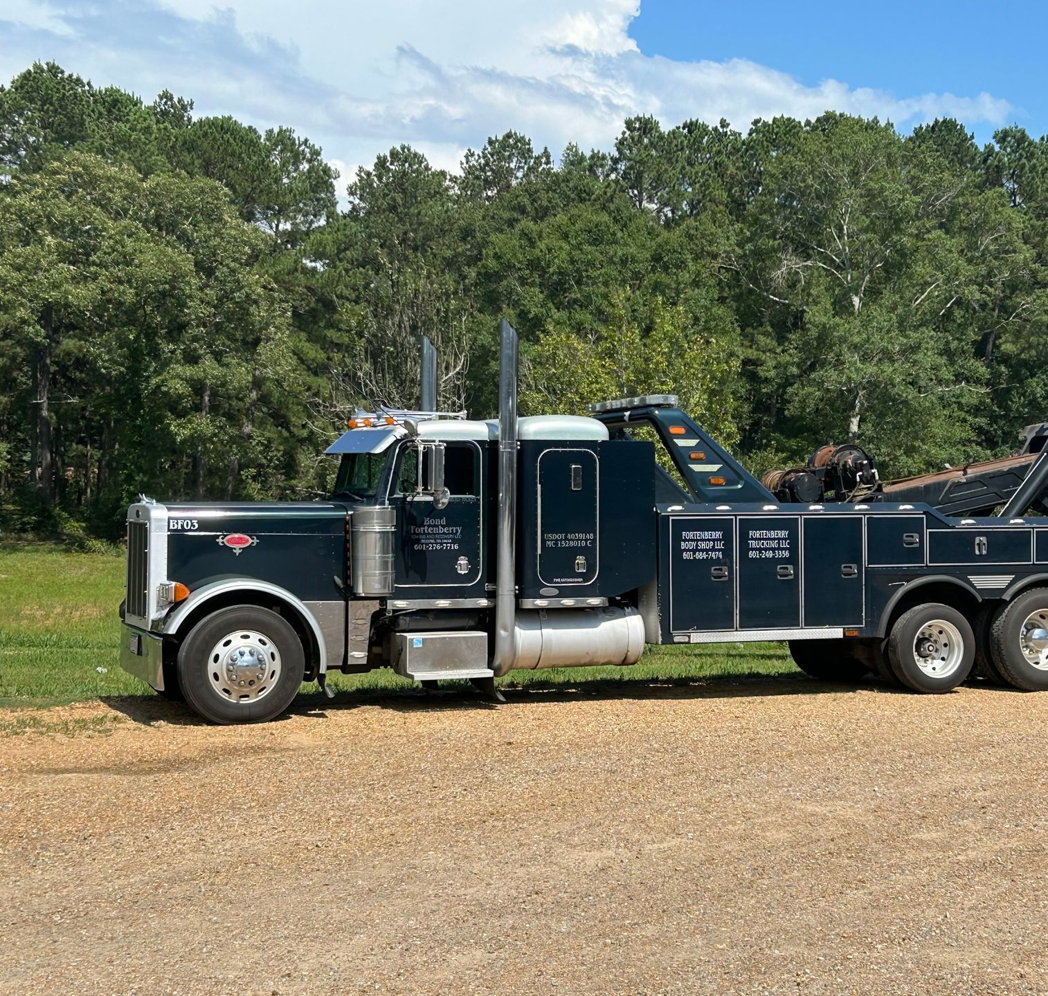 Black and silver tow truck parked on gravel road, trees in the background.