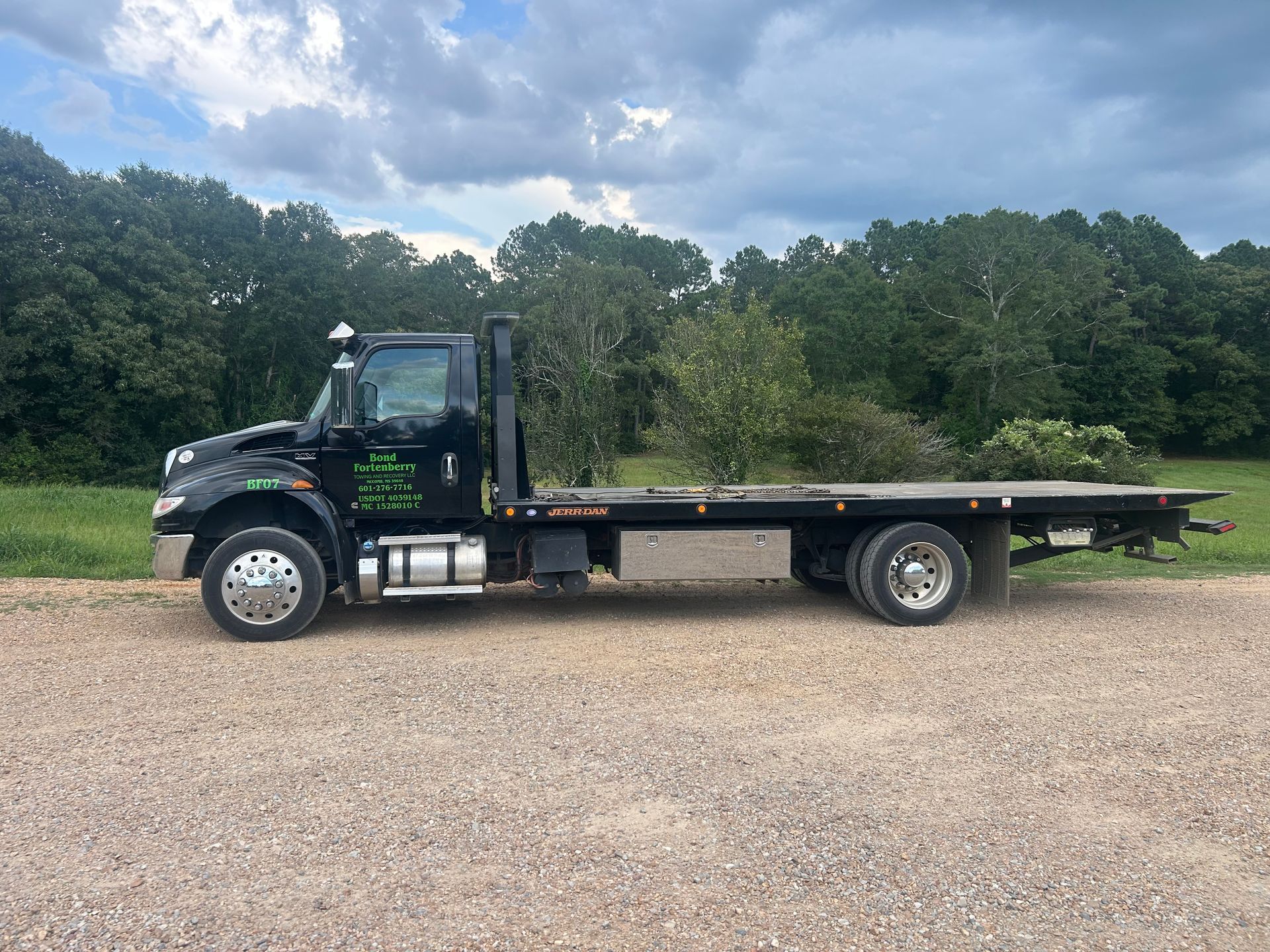 Black flatbed tow truck parked on gravel with trees in the background.