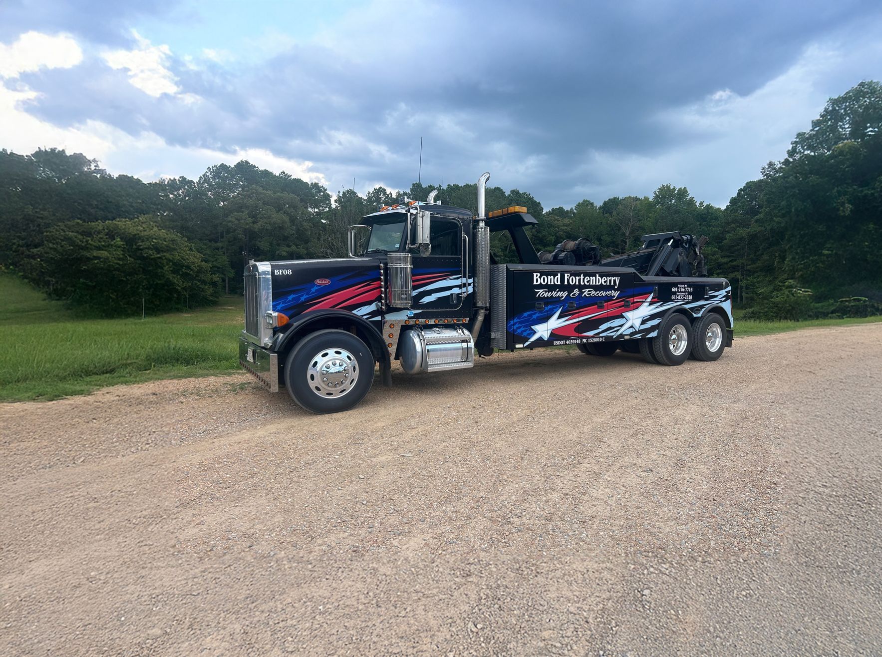 Tow truck, dark blue with red and white graphics, parked on gravel road, cloudy sky, trees.