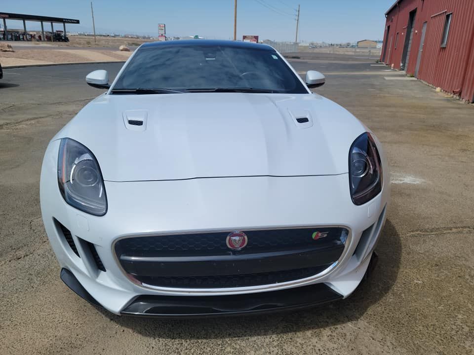 White Jaguar F-Type sports car, parked in front of a red building, with a black roof and a black front grill.