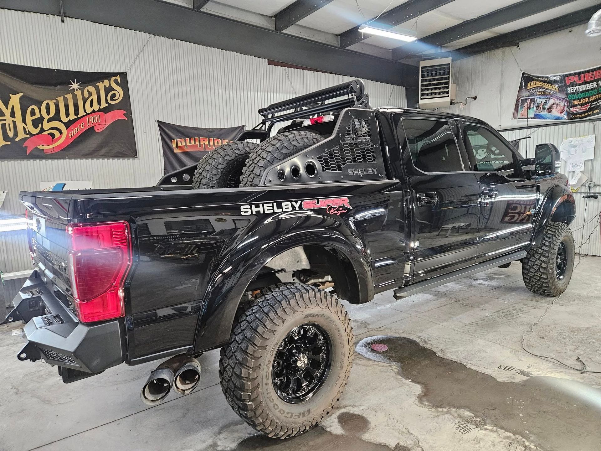 Black Shelby Super Truck with off-road tires, spare tires, and roof rack parked in a shop.