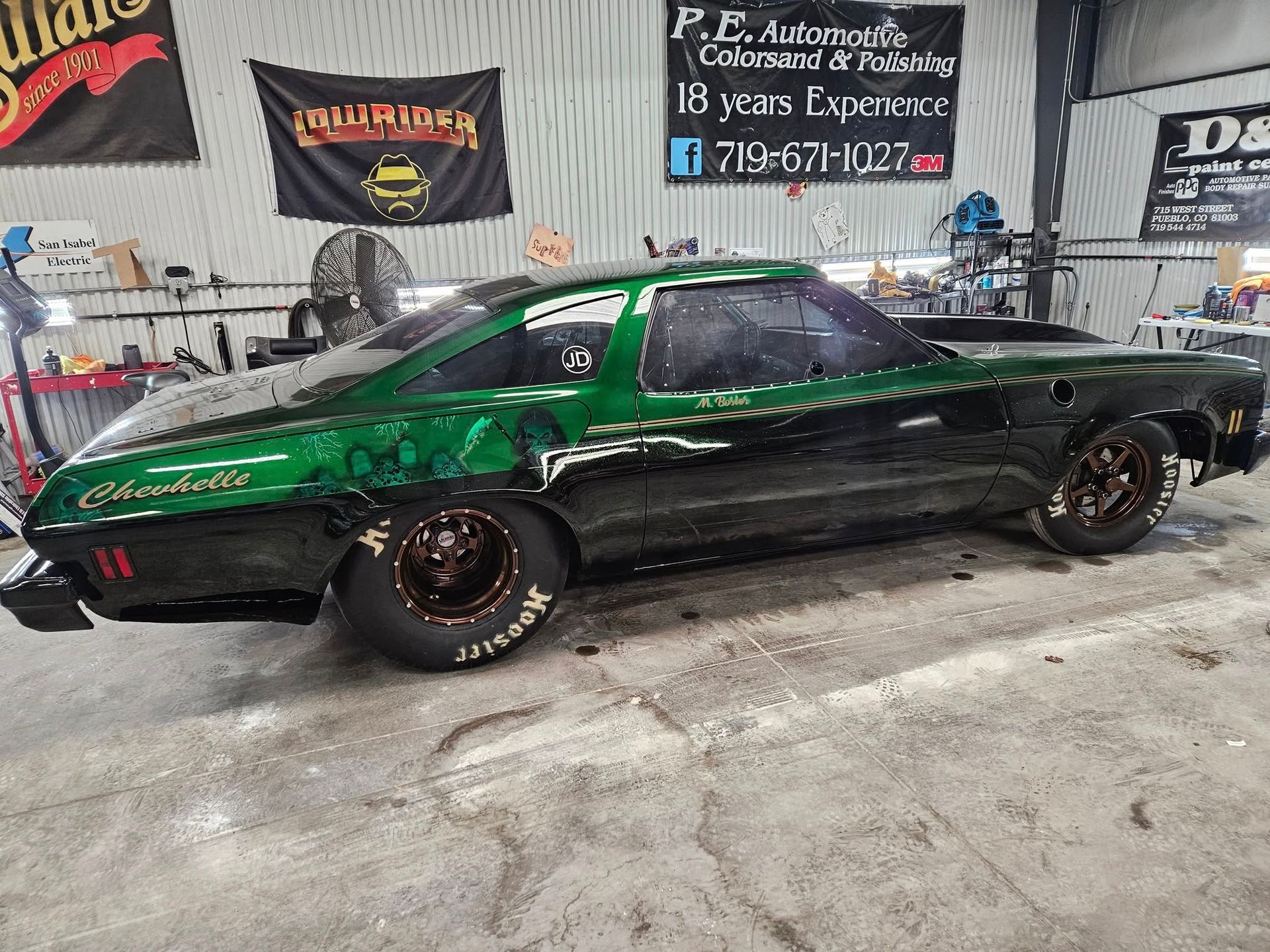 Green and black Chevrolet drag car in a garage, with sponsor signs in the background.