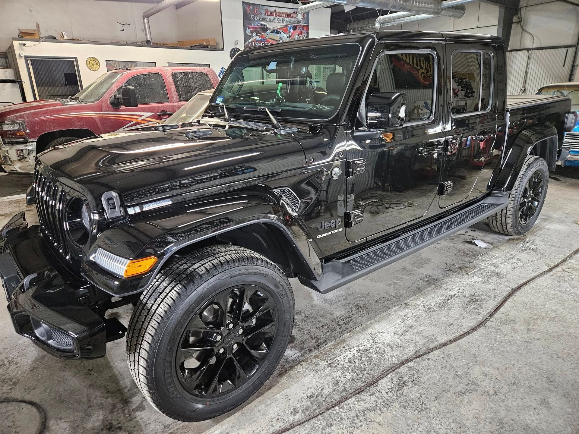 Black Jeep Gladiator pickup truck in a shop with a red truck in background.