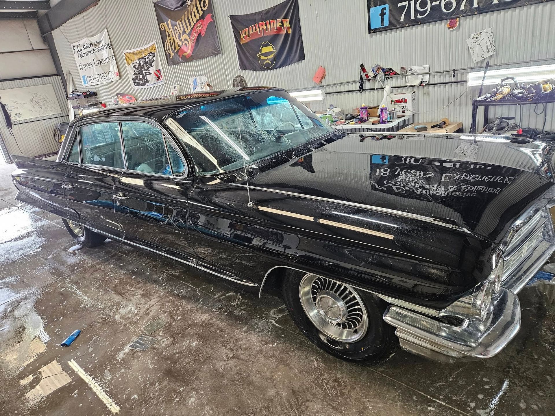 Black classic car, possibly a Cadillac, inside a garage with banners and other items in the background.