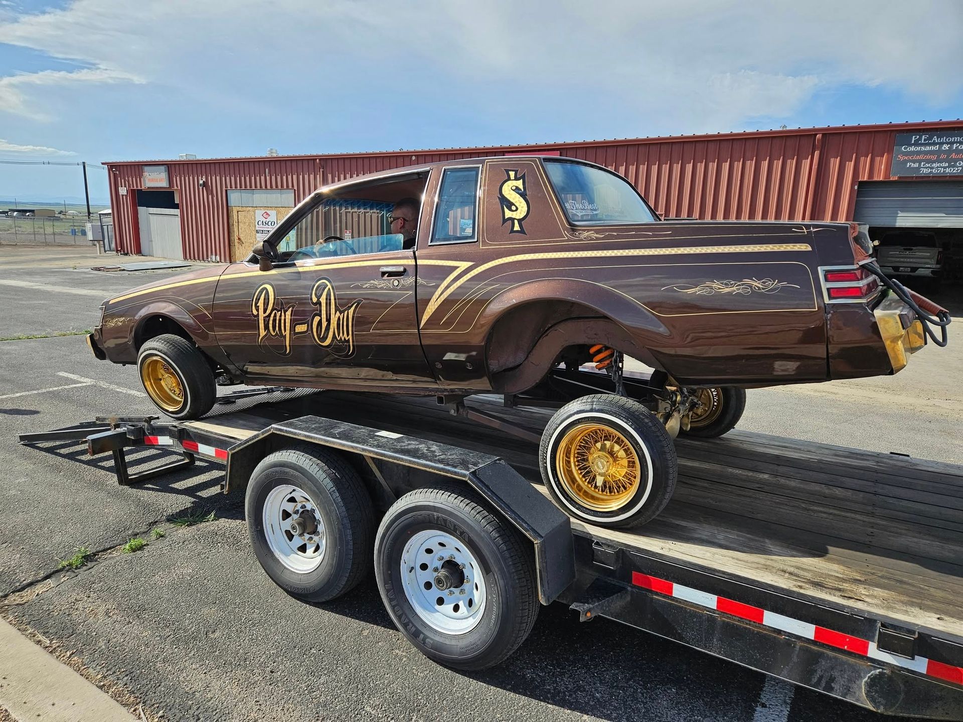 Brown lowrider car on a trailer, gold wheels, brown paint with gold accents, outdoor setting.