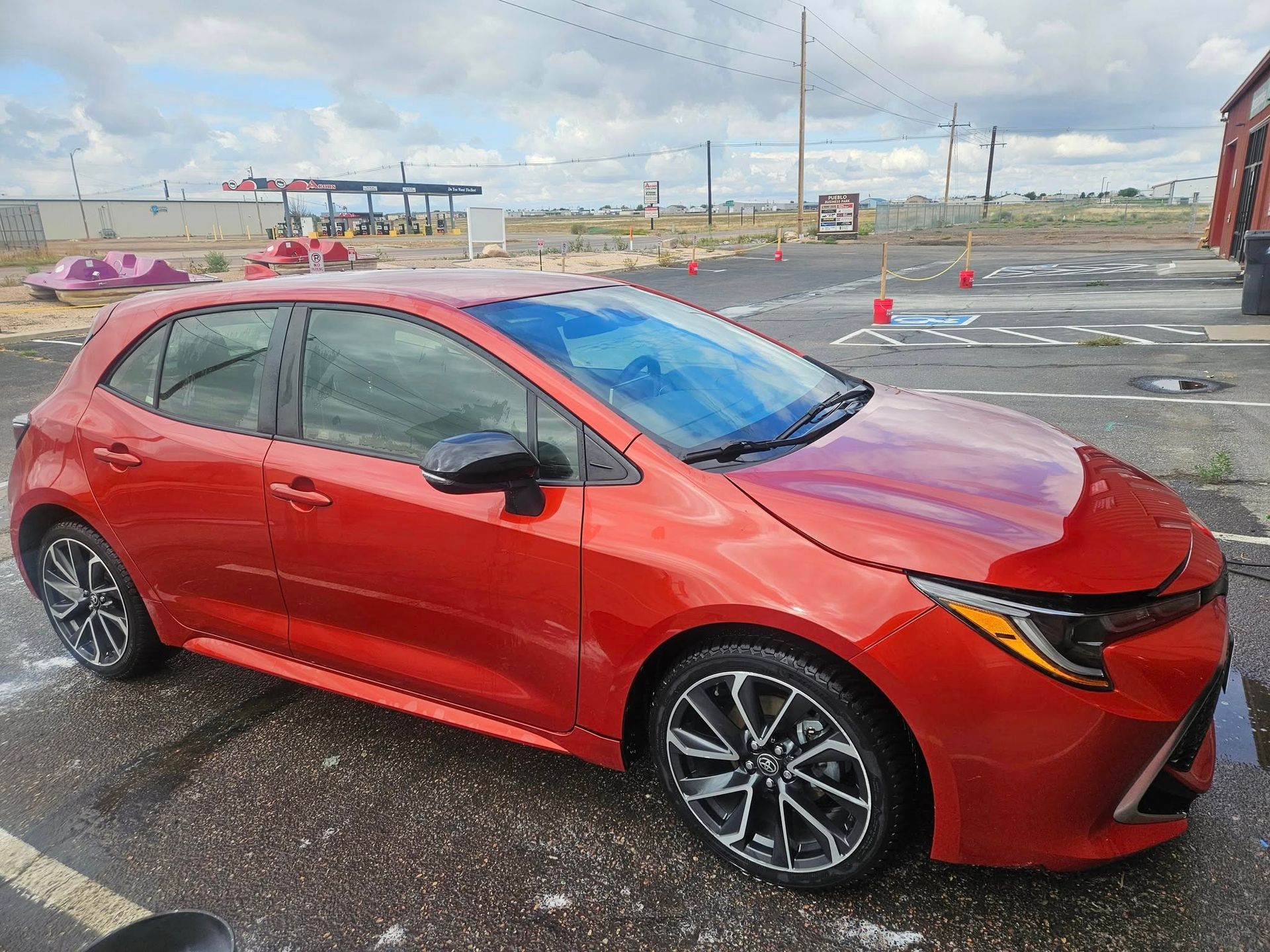 Red Toyota Corolla hatchback parked on a paved lot; gas station and sky visible in the background.