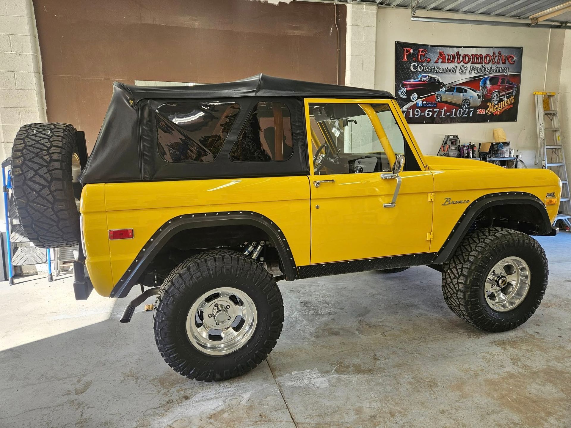 Yellow vintage Ford Bronco with a black soft top, spare tire, and large off-road tires.