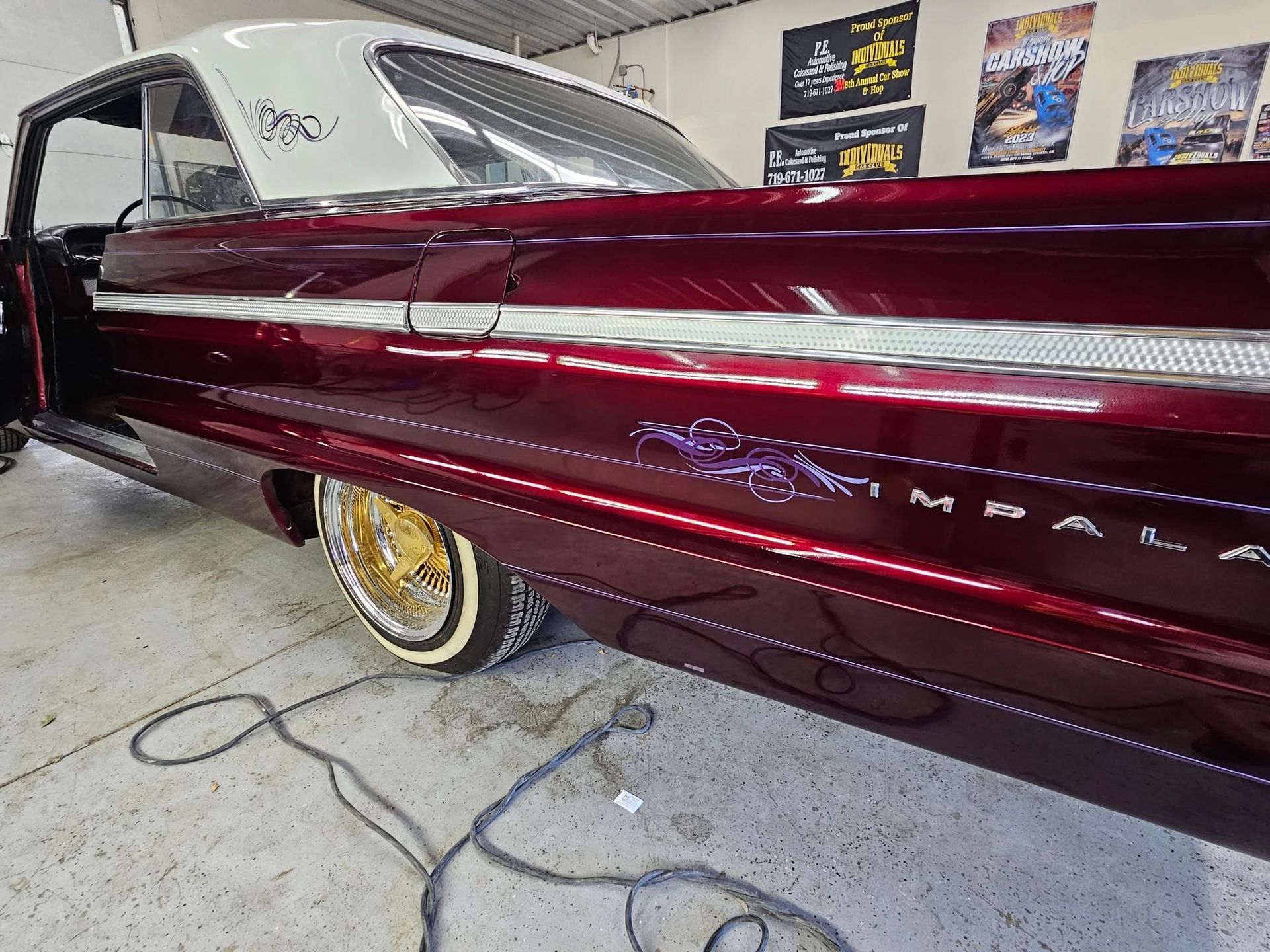 Burgundy lowrider car with gold rims and white roof, indoors. Features custom paint and trim.