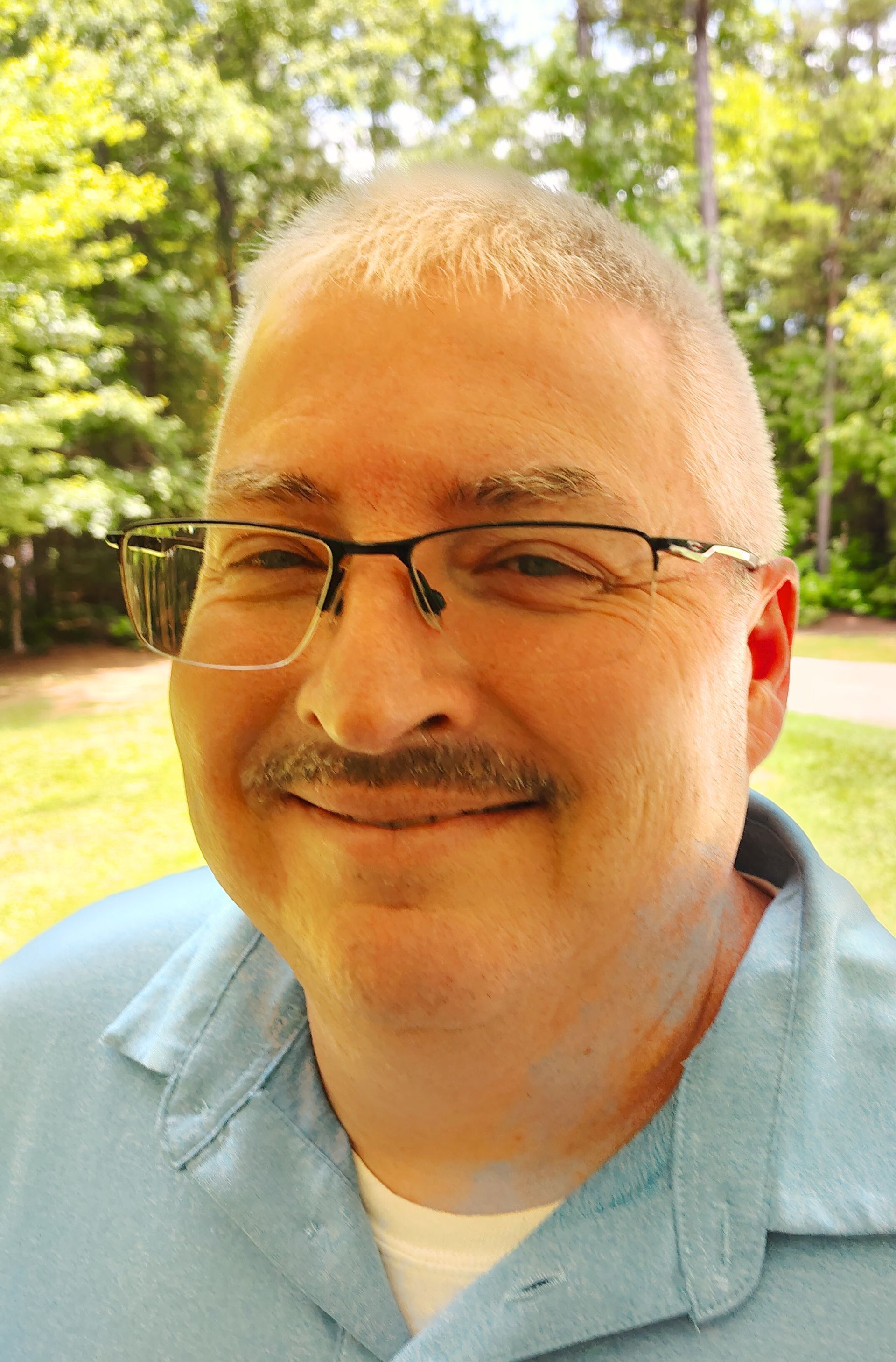 A person wearing glasses and a light blue polo shirt, smiling, with a blurred green park background.