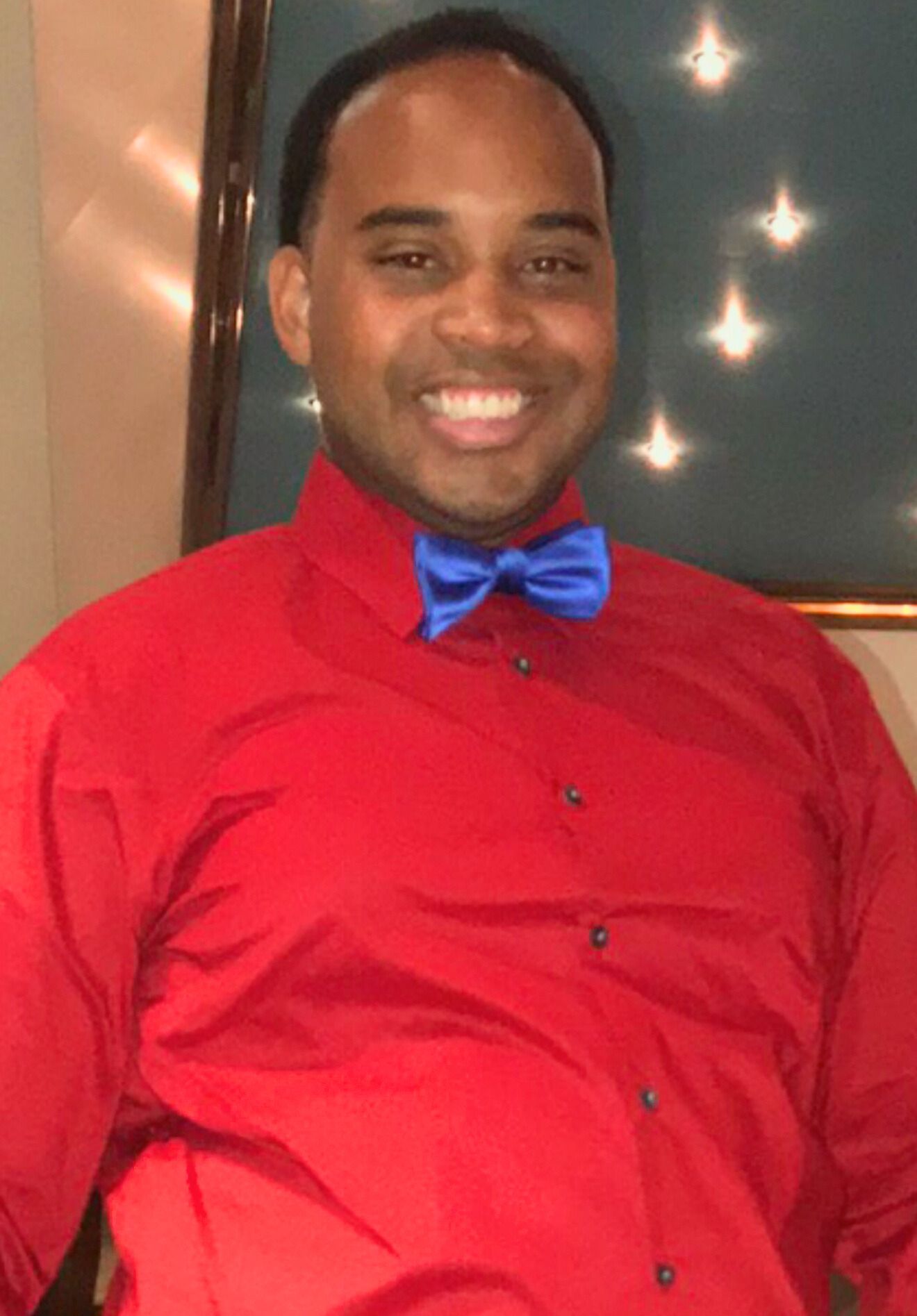 A smiling person wearing a red button-down shirt and a bright blue bow tie, indoors in front of a framed dark-blue panel.