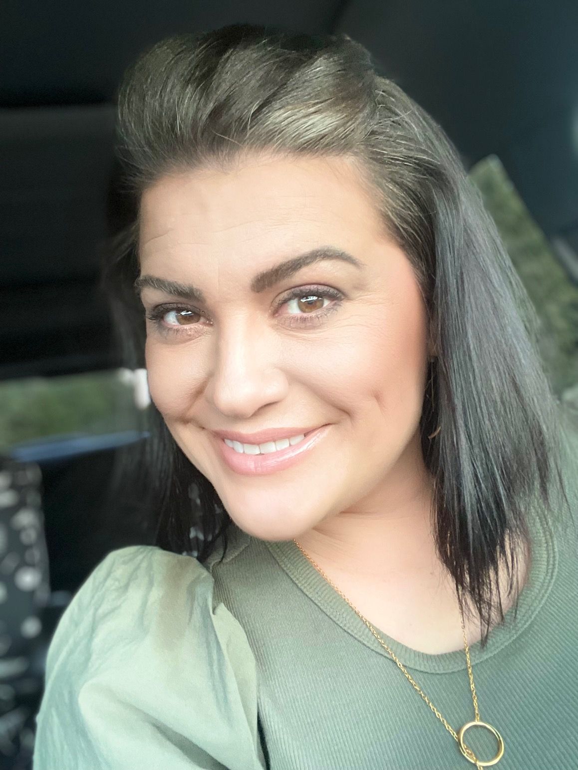 A smiling person with dark hair styled back, wearing a green top and a gold pendant necklace.