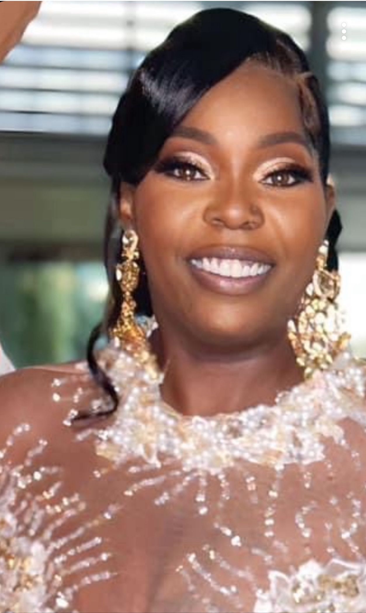A smiling person with dark hair, wearing large gold statement earrings and a beaded, semi-sheer white garment.