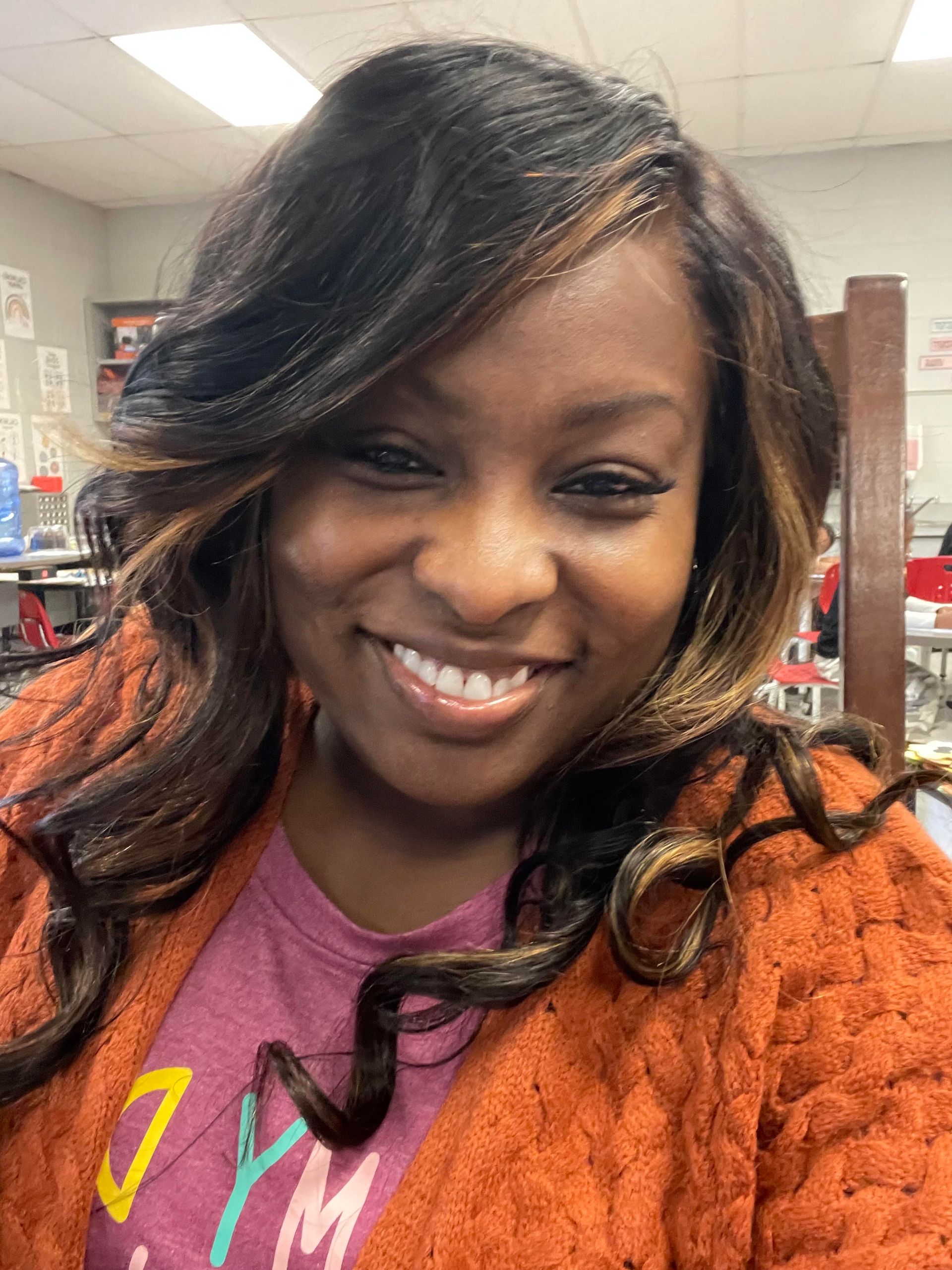 A smiling person with long, dark, wavy hair with highlights, wearing an orange cardigan over a purple shirt.