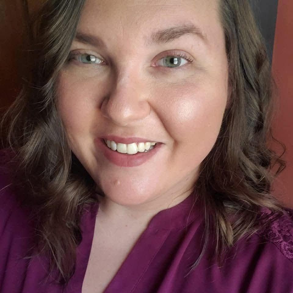 A person with shoulder-length wavy brown hair smiling at the camera, wearing a deep purple top against a dark background.
