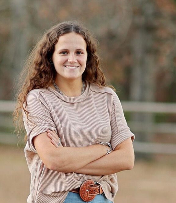 A smiling person with long wavy hair, wearing a ribbed tan top and a decorative belt, posing with arms crossed outdoors.