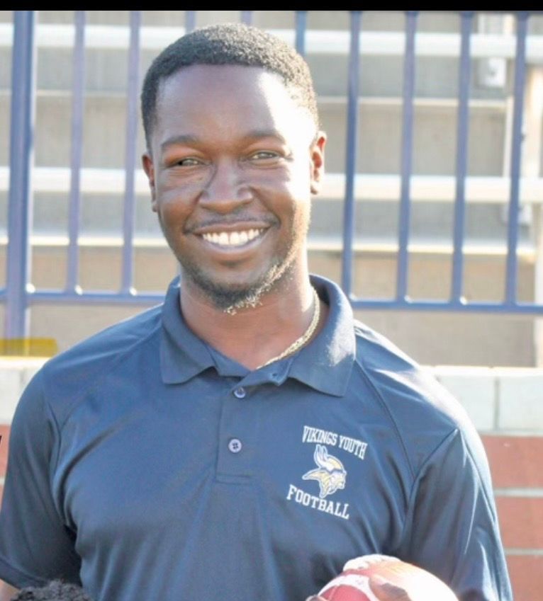 Smiling coach wearing a navy blue Vikings Youth Football polo shirt while standing in front of stadium seating.