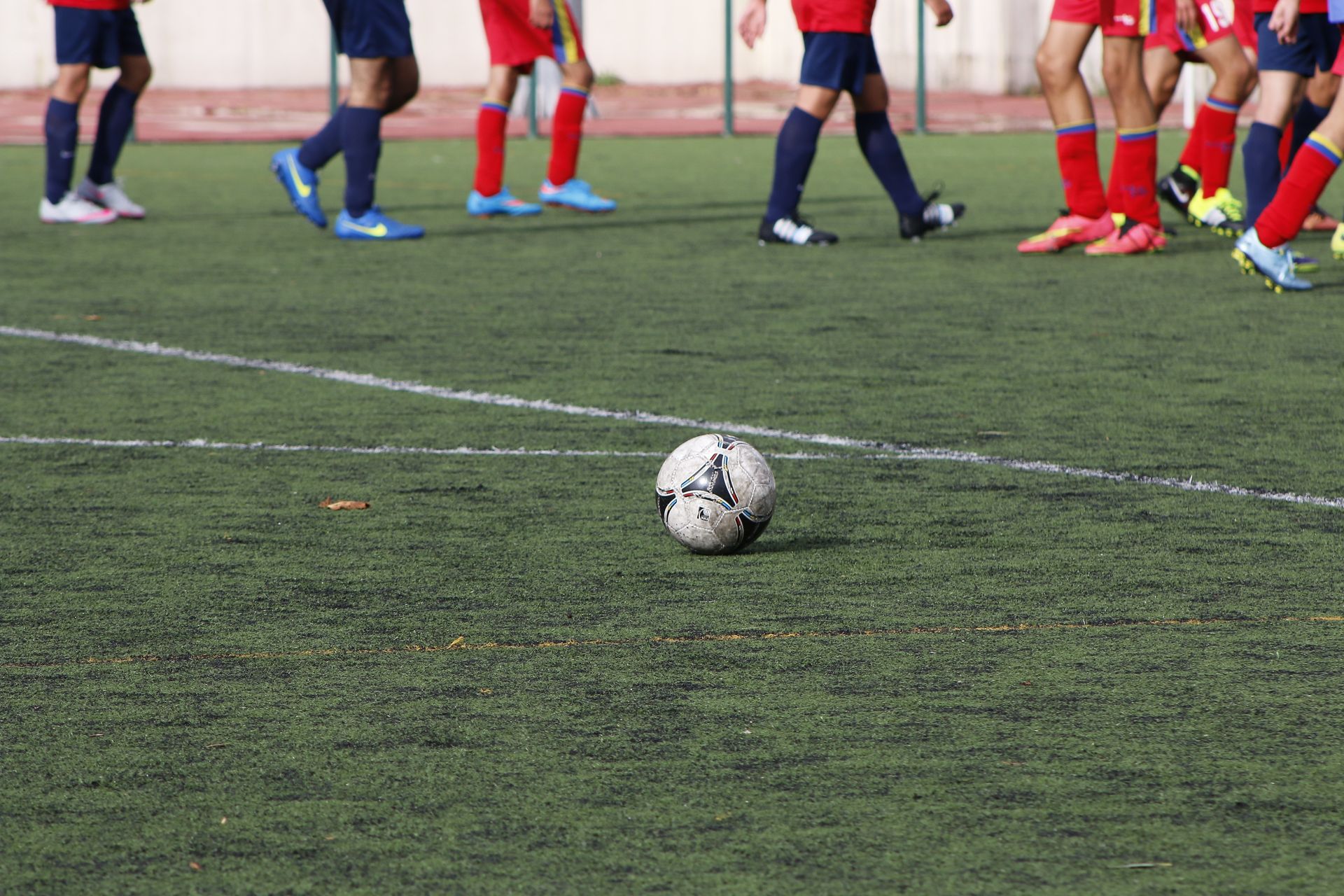 A soccer ball sits on an artificial turf field, with a group of players in red jerseys and blue socks standing behind it.