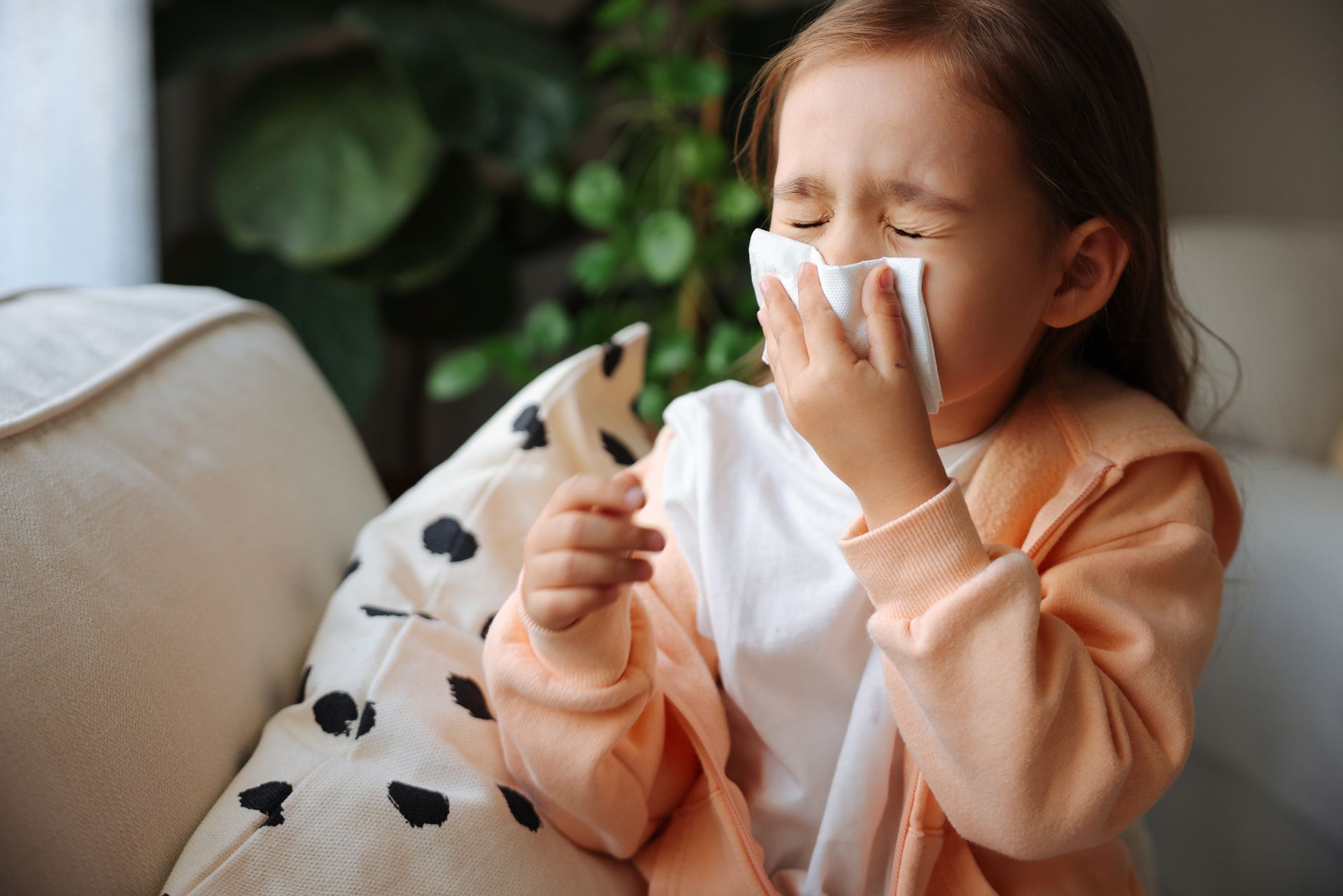 A little girl is blowing her nose into a napkin while sitting on a couch.