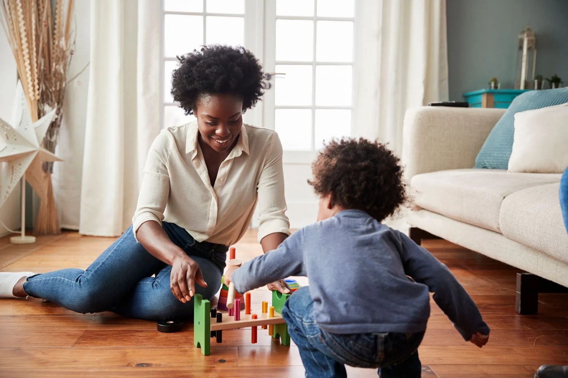 An adult and a child play with a colorful wooden toy on a wooden floor in a sunlit living room.