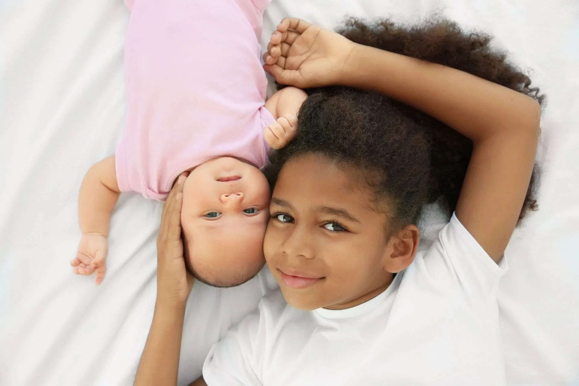 A child wearing a white shirt lies on a white surface, gently holding a baby in a pink shirt next to their head.
