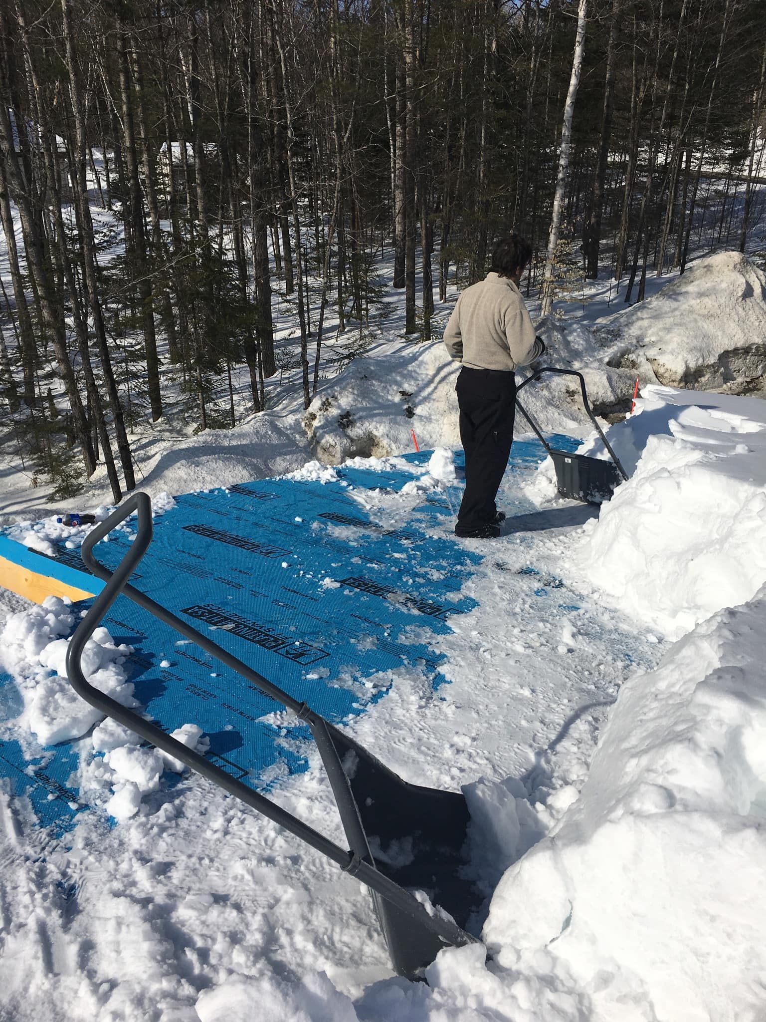 Une personne pellete la neige d'une bâche bleue dans les bois.