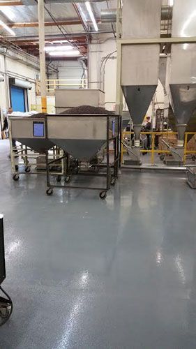 Factory interior with industrial machinery. A large bin holds dark material, near metal silos and a worker.