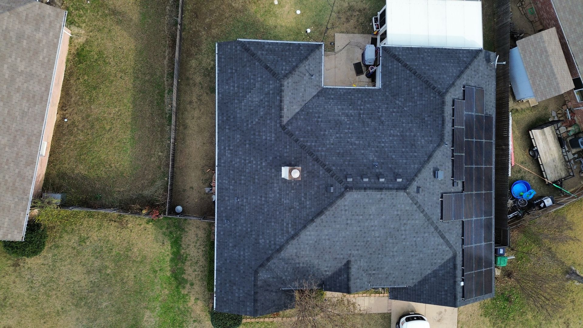 Overhead view of a dark gray house roof with solar panels, surrounded by grass and adjacent houses.
