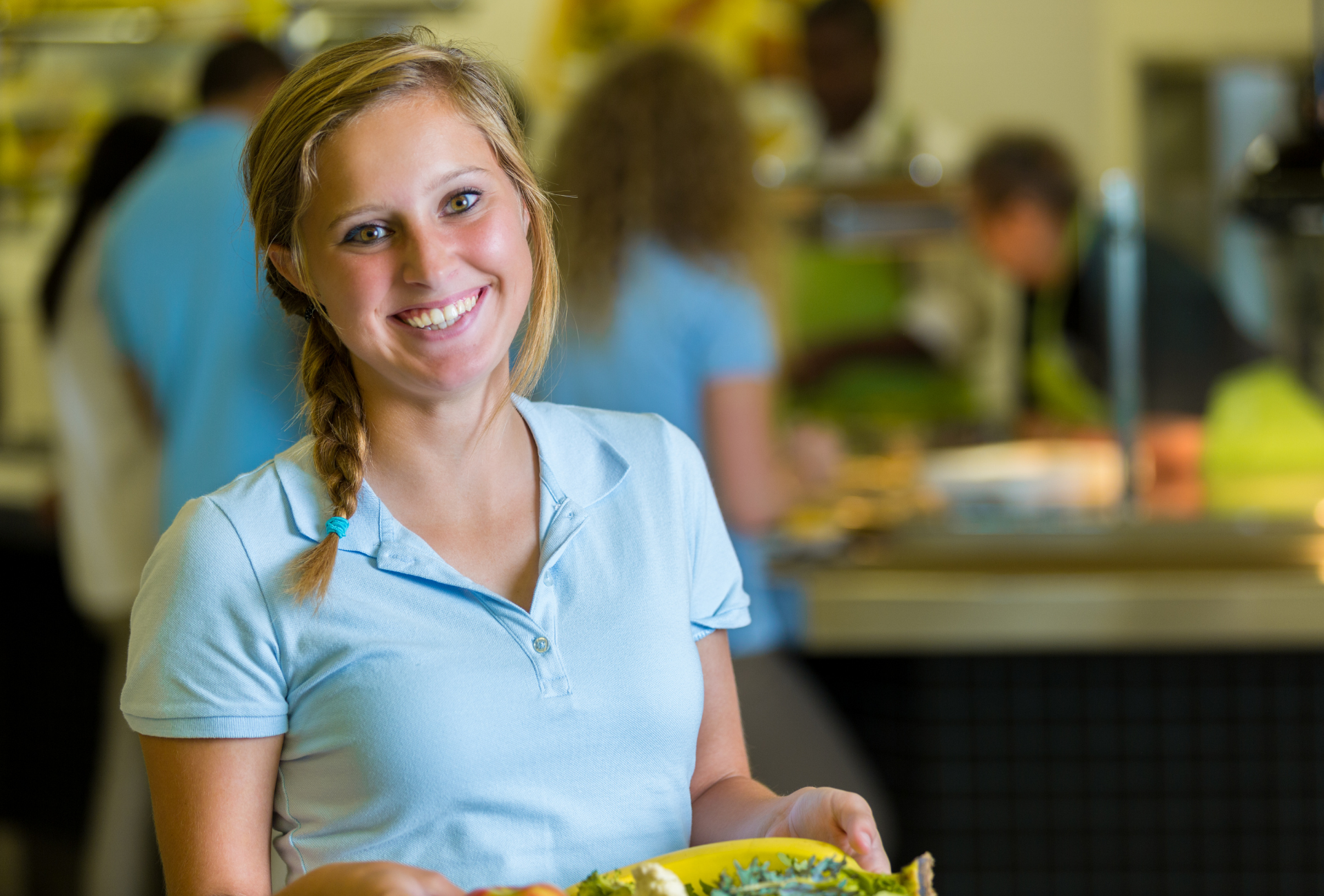 Smiling woman holding a tray with food in a cafeteria. Wearing light blue shirt; blonde hair in braid.