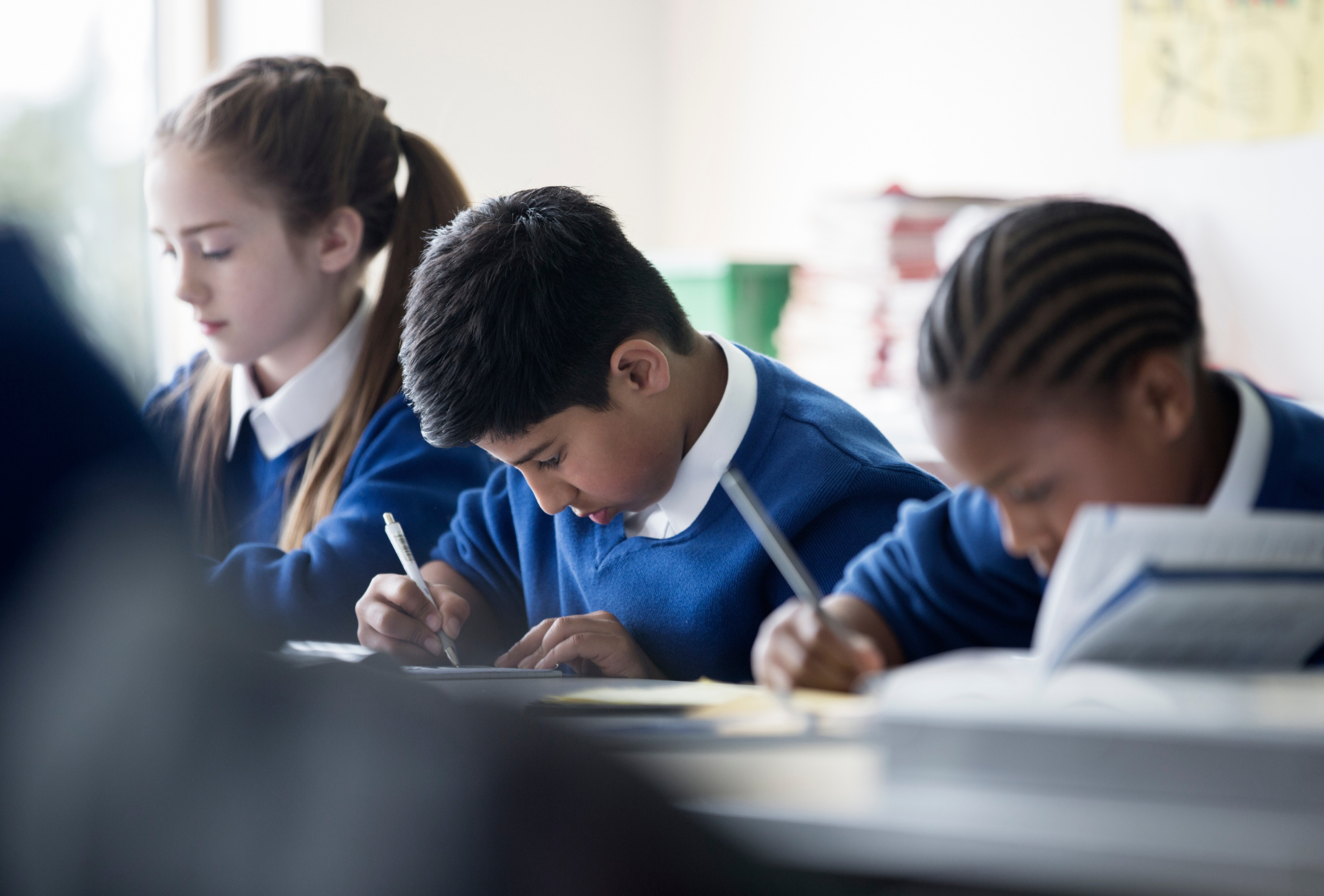 Three children in blue uniforms writing at desks in a classroom.