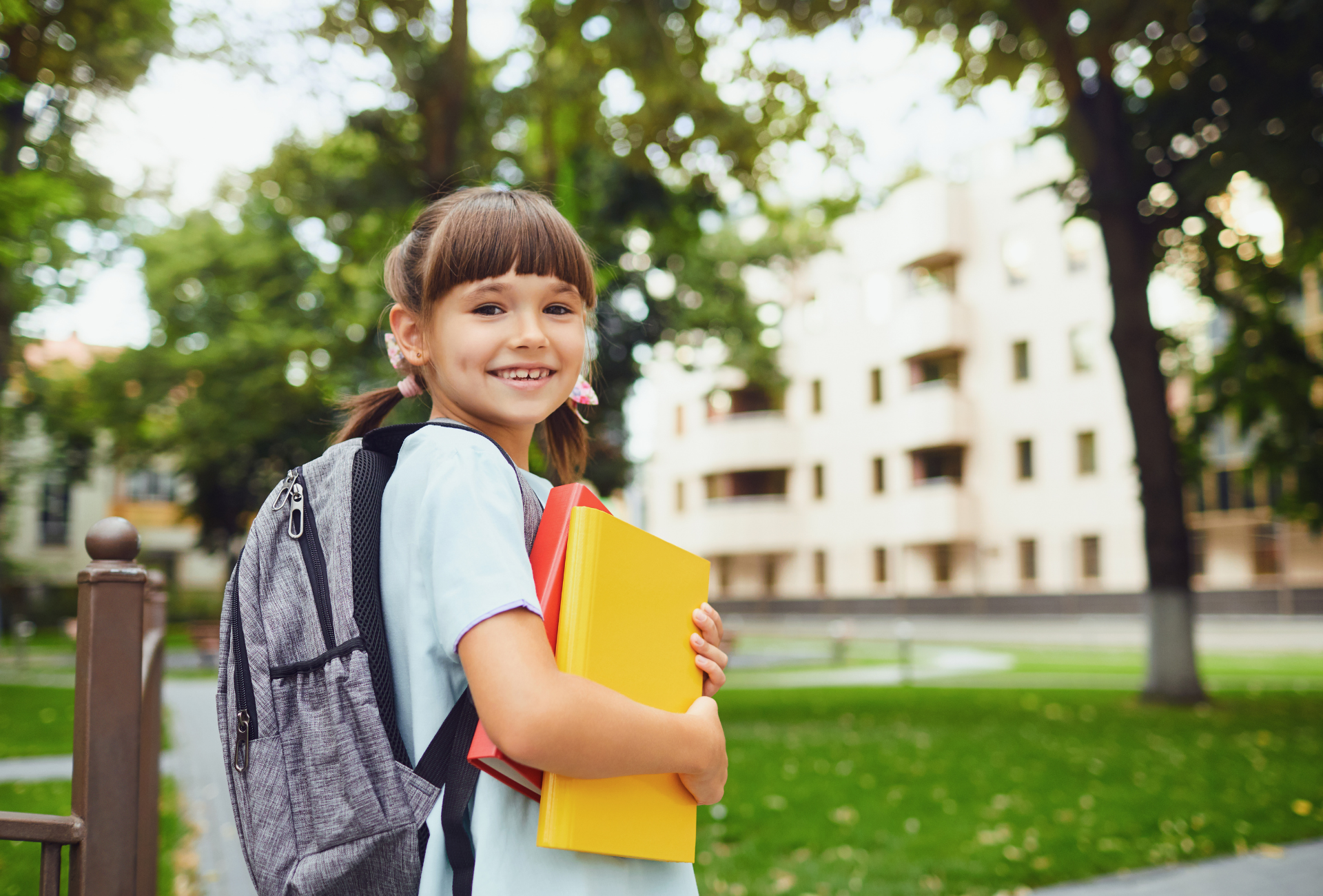 Girl with backpack and books smiles in front of a school building.