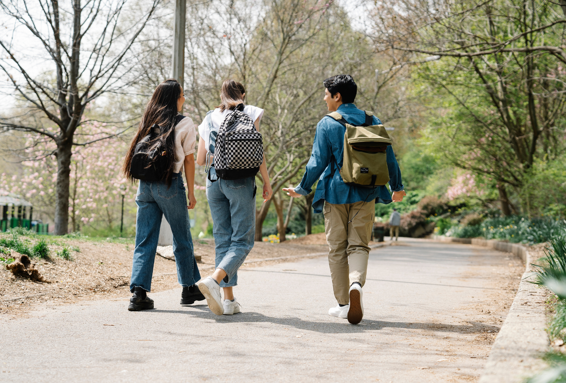 Three young people with backpacks walk on a path in a park, trees and greenery surround them.