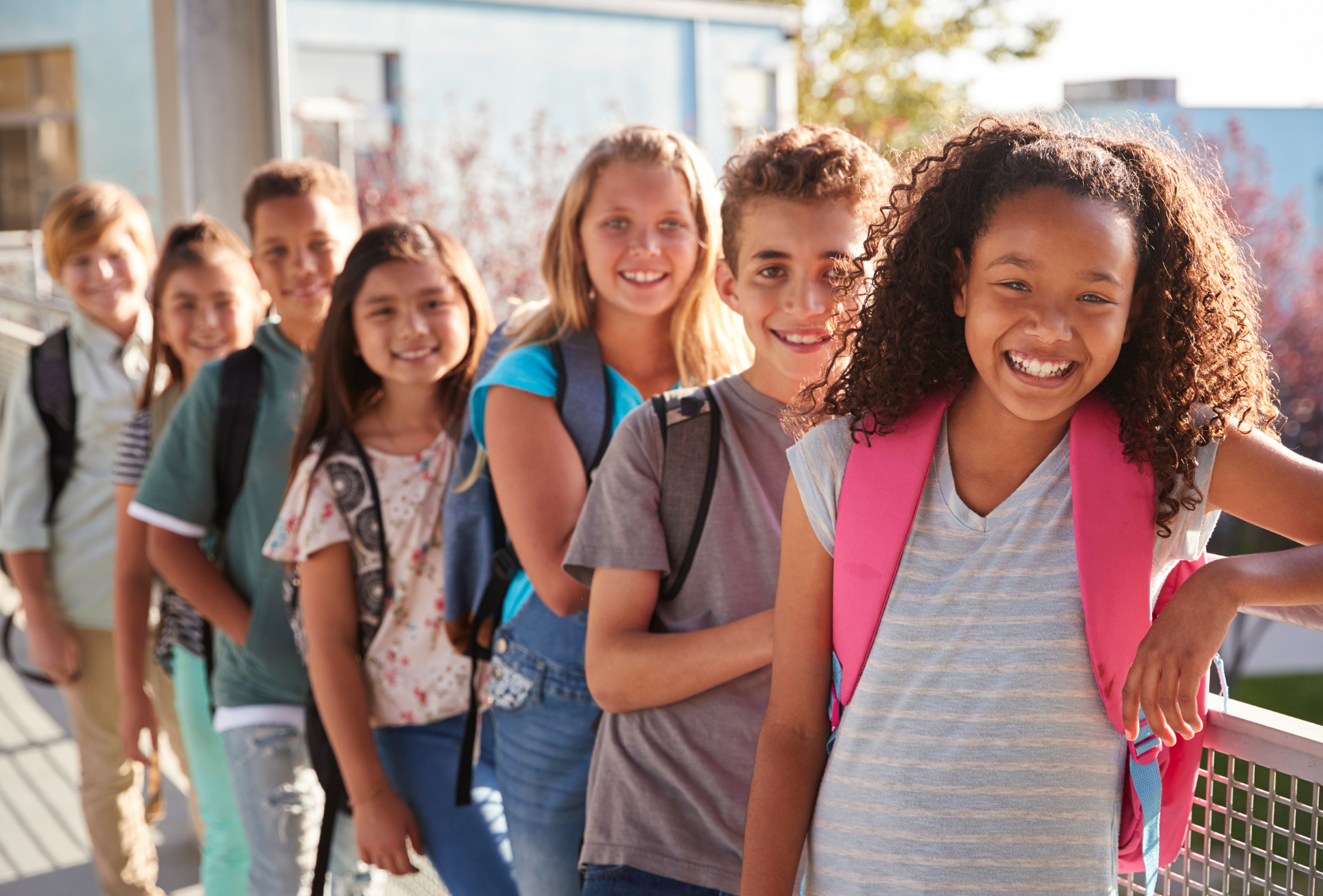 Group of diverse schoolchildren smiling, wearing backpacks, standing in a line outdoors.