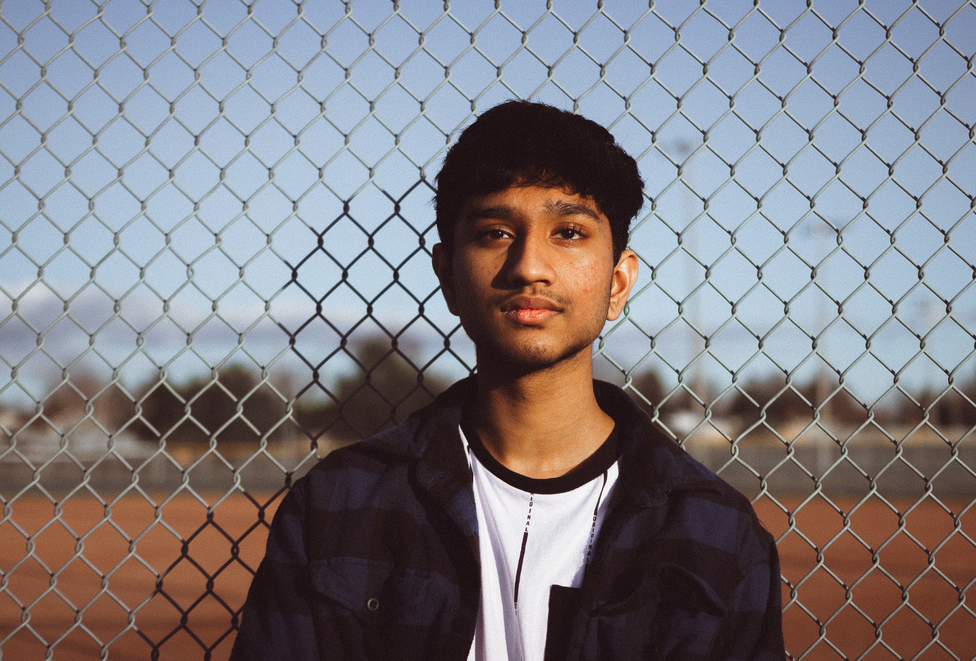 Young man in front of a chain-link fence, wearing a flannel shirt.