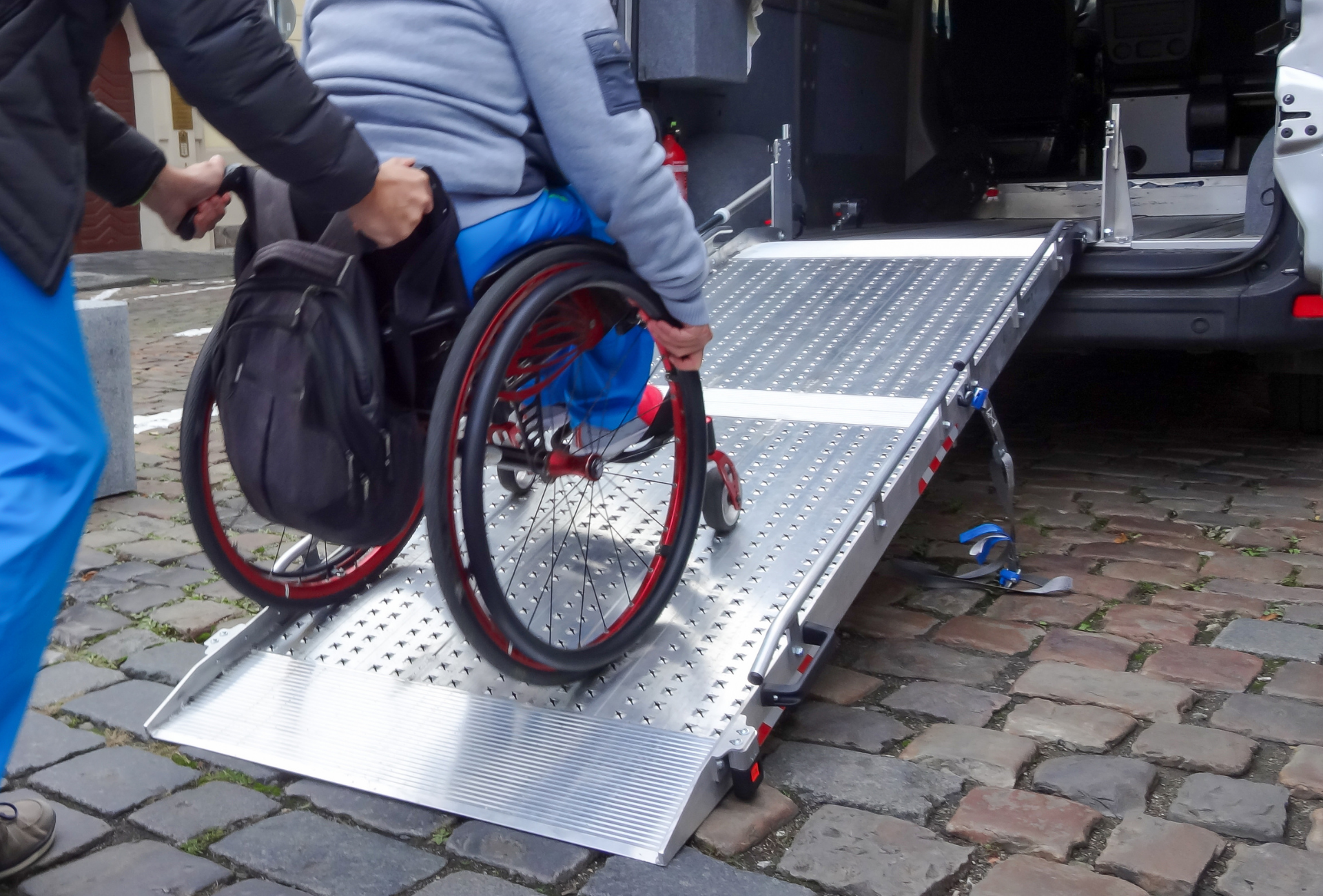Person in wheelchair assisted onto a ramp into a van by another person, cobblestone street.