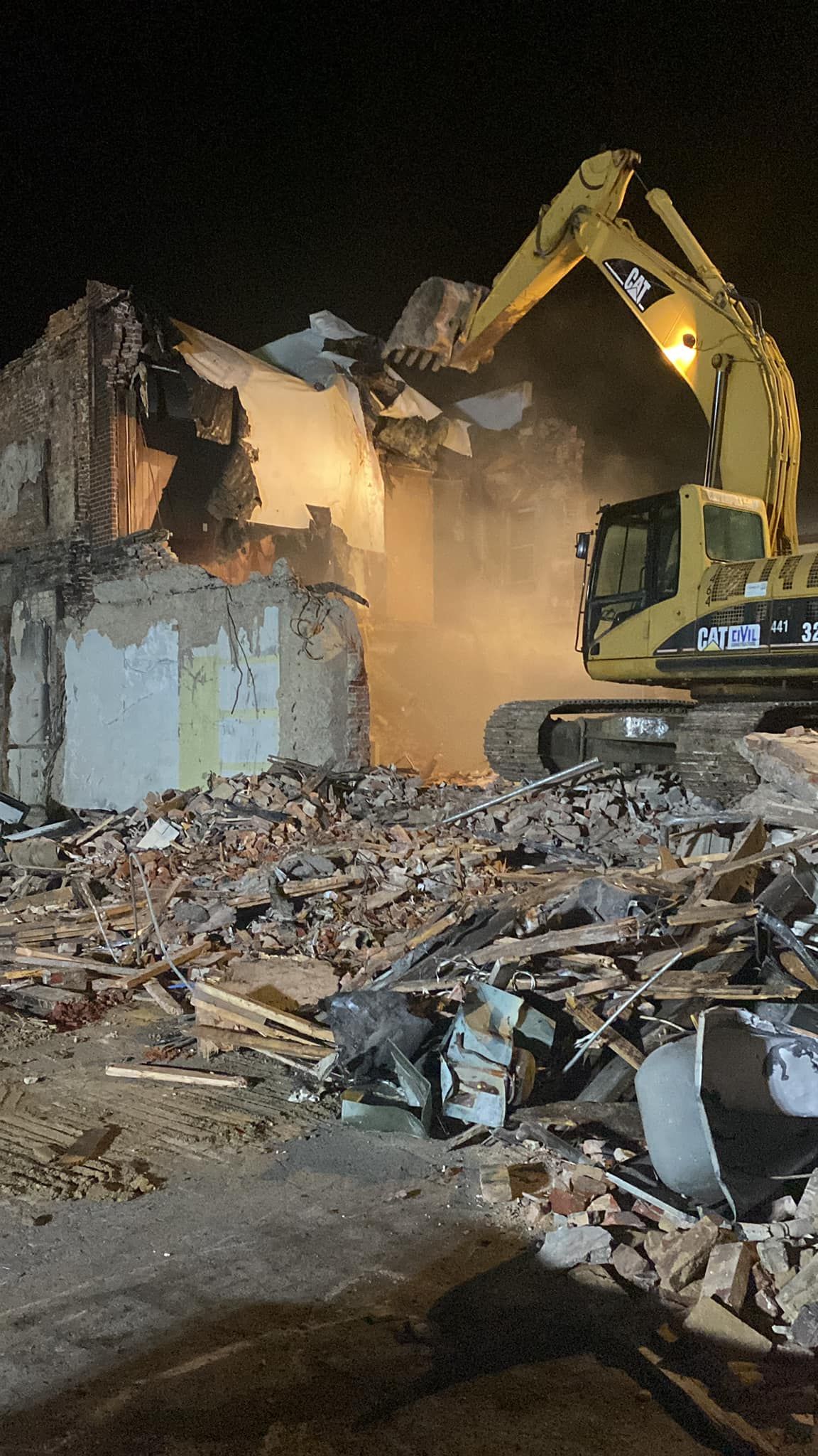 A yellow excavator demolishes a damaged building amid a pile of rubble at night.