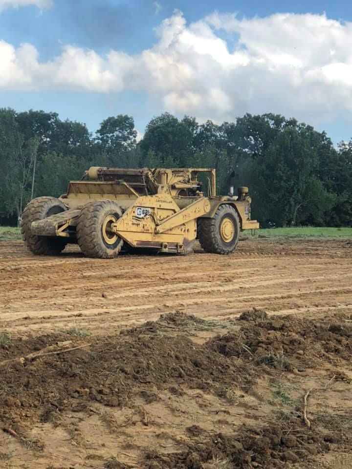 A worker in a hard hat stands on the tracks of a large yellow excavator while performing maintenance at a rocky site.