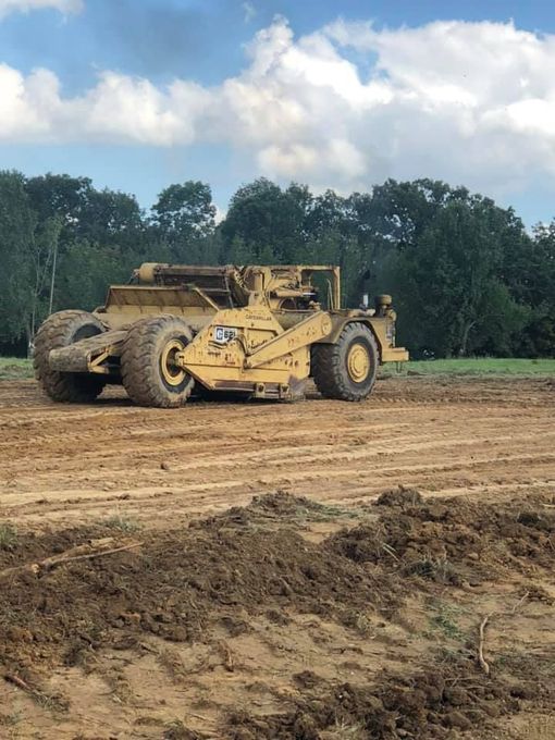 A worker in a hard hat stands on the tracks of a large yellow excavator while performing maintenance at a rocky site.
