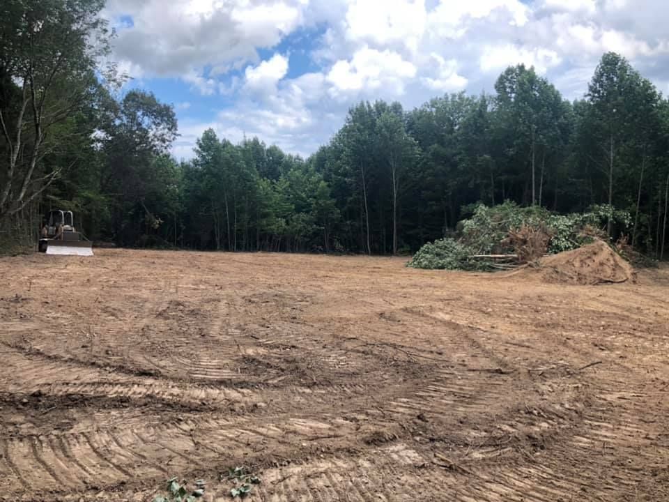 A cleared, dirt construction site with tire tracks, a pile of brush, and a dense tree line under a cloudy sky.