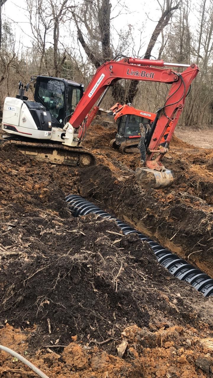 A red and white excavator digs a trench in a wooded area, with a long, black corrugated drainage pipe laid inside.