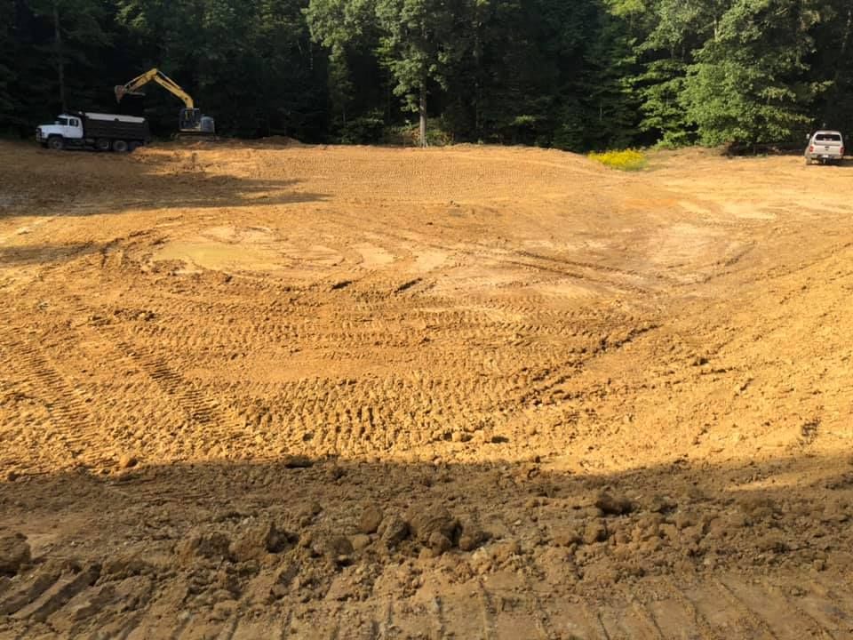 An excavator and a dump truck work on a large, dirt-filled construction site surrounded by trees.