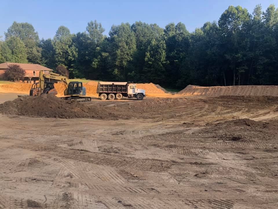 An excavator loads dirt into a dump truck at a construction site with a treeline in the background.