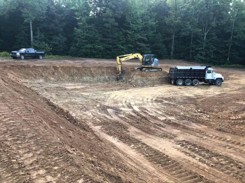 An excavator loads soil into a white dump truck at an active construction site near a tree line.