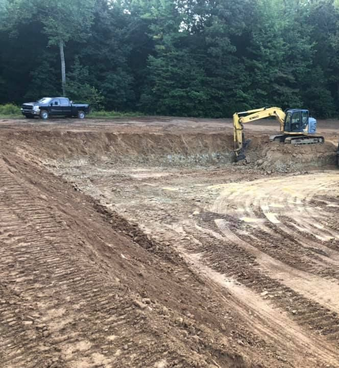 A yellow excavator loads dirt into a dump truck at a wooded construction site with a black pickup truck nearby.