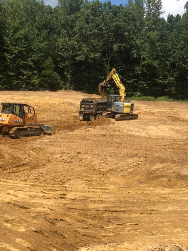 A yellow excavator loads dirt into a dump truck on a large, cleared construction site near a dense forest.