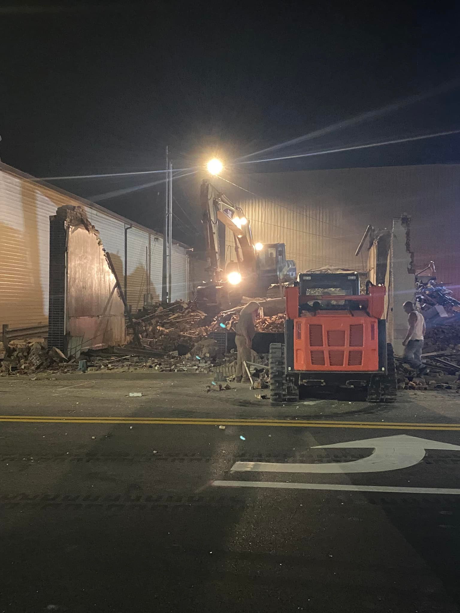 At night, a red skid steer loader works on the demolition of a building structure, surrounded by debris and construction.
