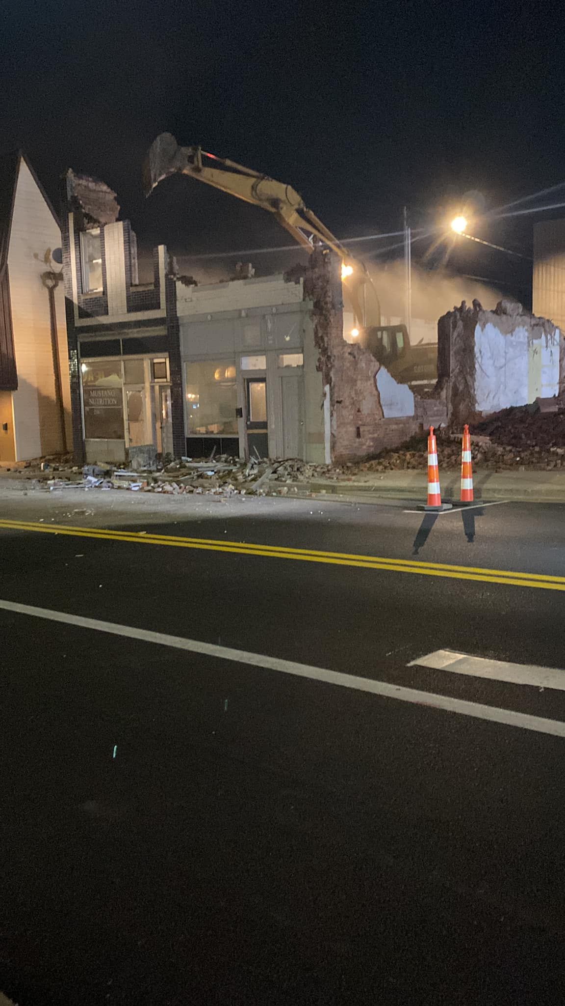 A yellow excavator demolishes a building at night, with debris scattered near a road marked by orange traffic cones.