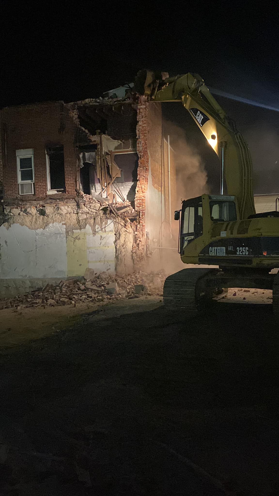 A large yellow excavator demolishes the brick wall of a building at night.