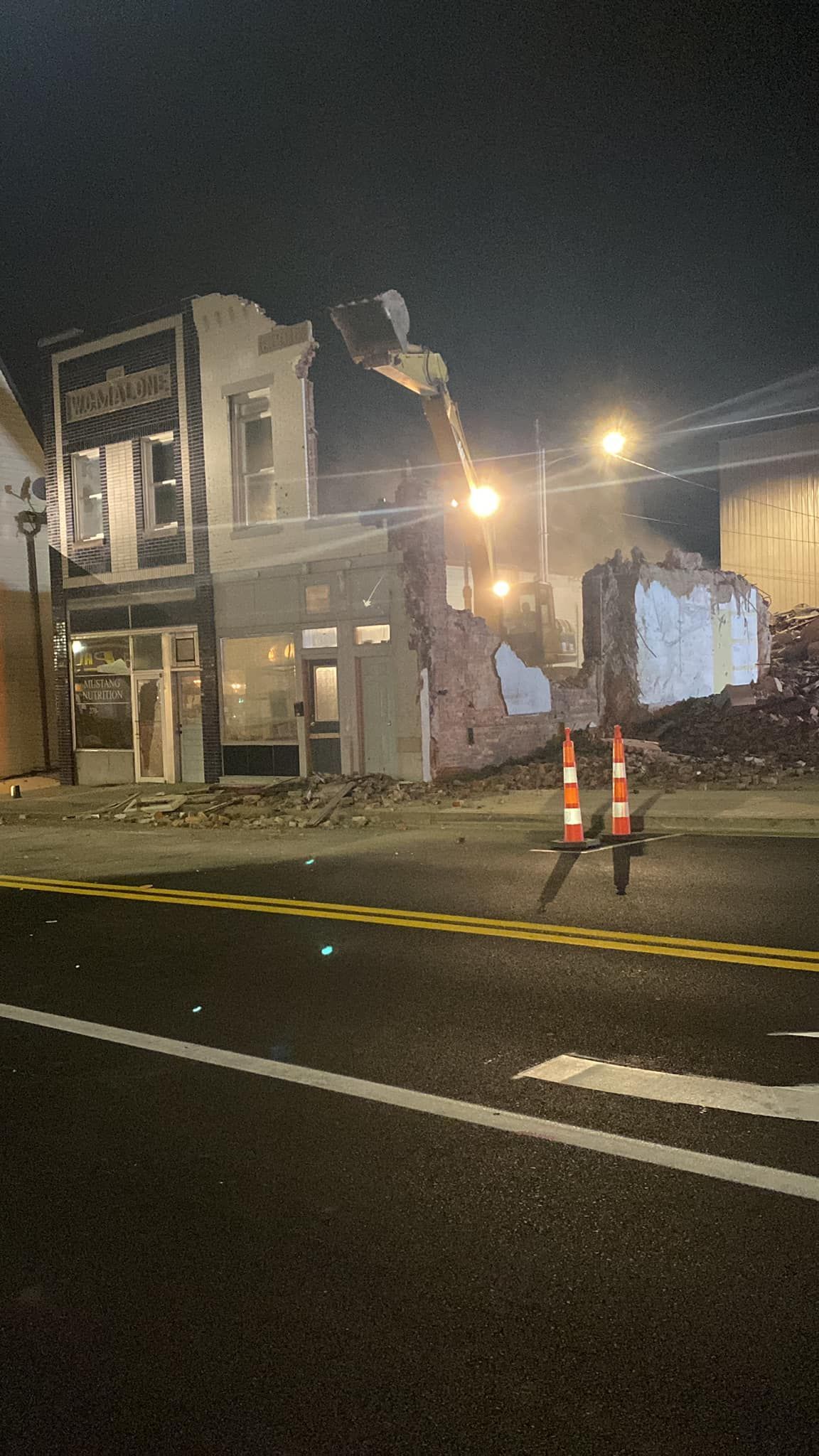A construction vehicle demolishes a building at night, with rubble in the foreground and traffic cones on the street.